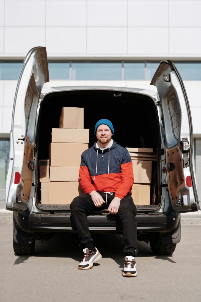 Man in a beanie and jacket sitting on a van tailgate surrounded by cardboard boxes.